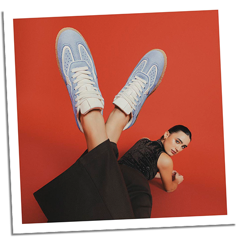 Mujer acostada usando tenis azules de moda y pantalón marrón en fondo rojo vibrante.