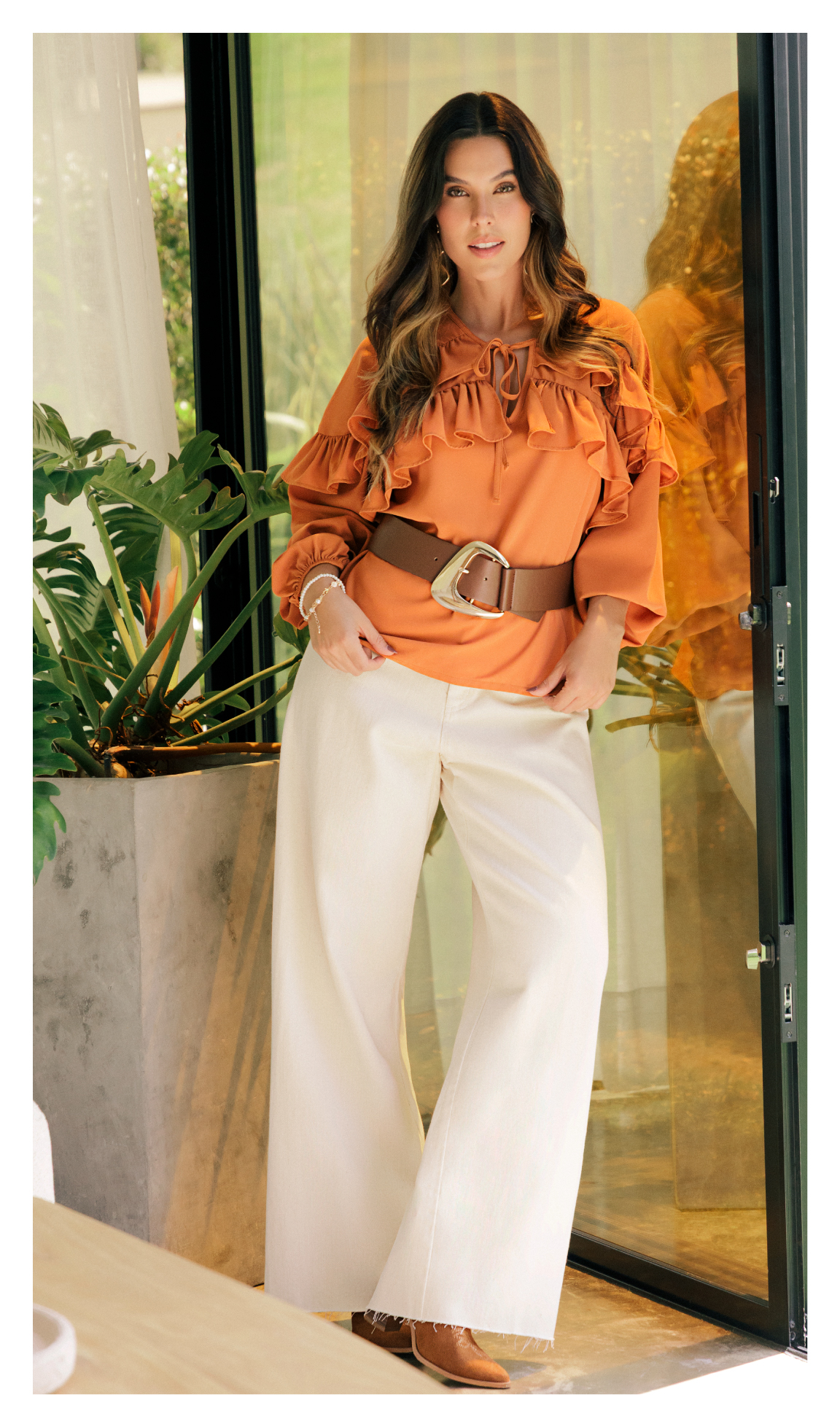 Mujer luciendo blusa naranja con boleros, maxi cinturón café y pantalón blanco.