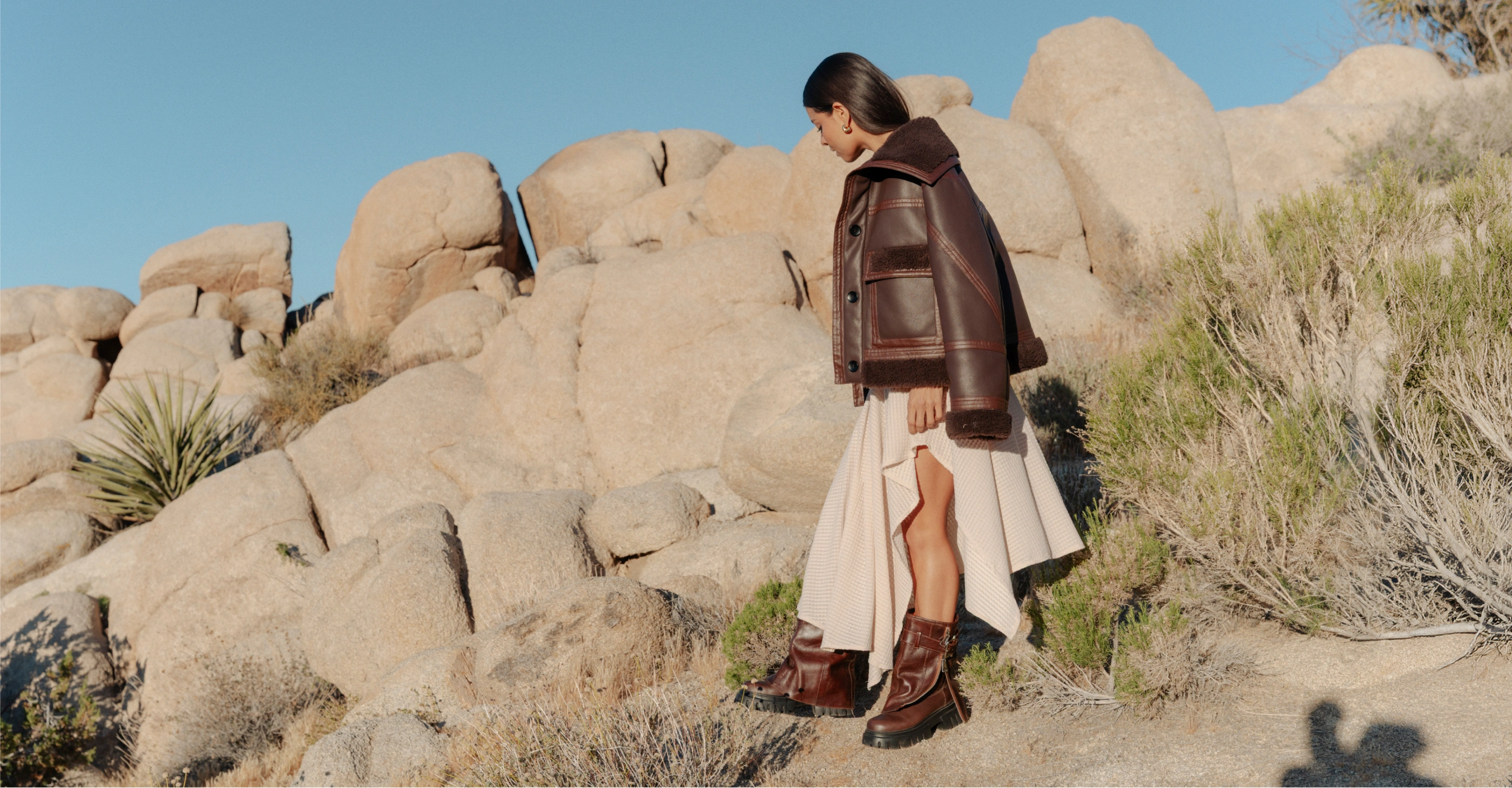 Mujer con chaqueta corta café, falda midi asimétrica beige y botas en un paisaje desértico.