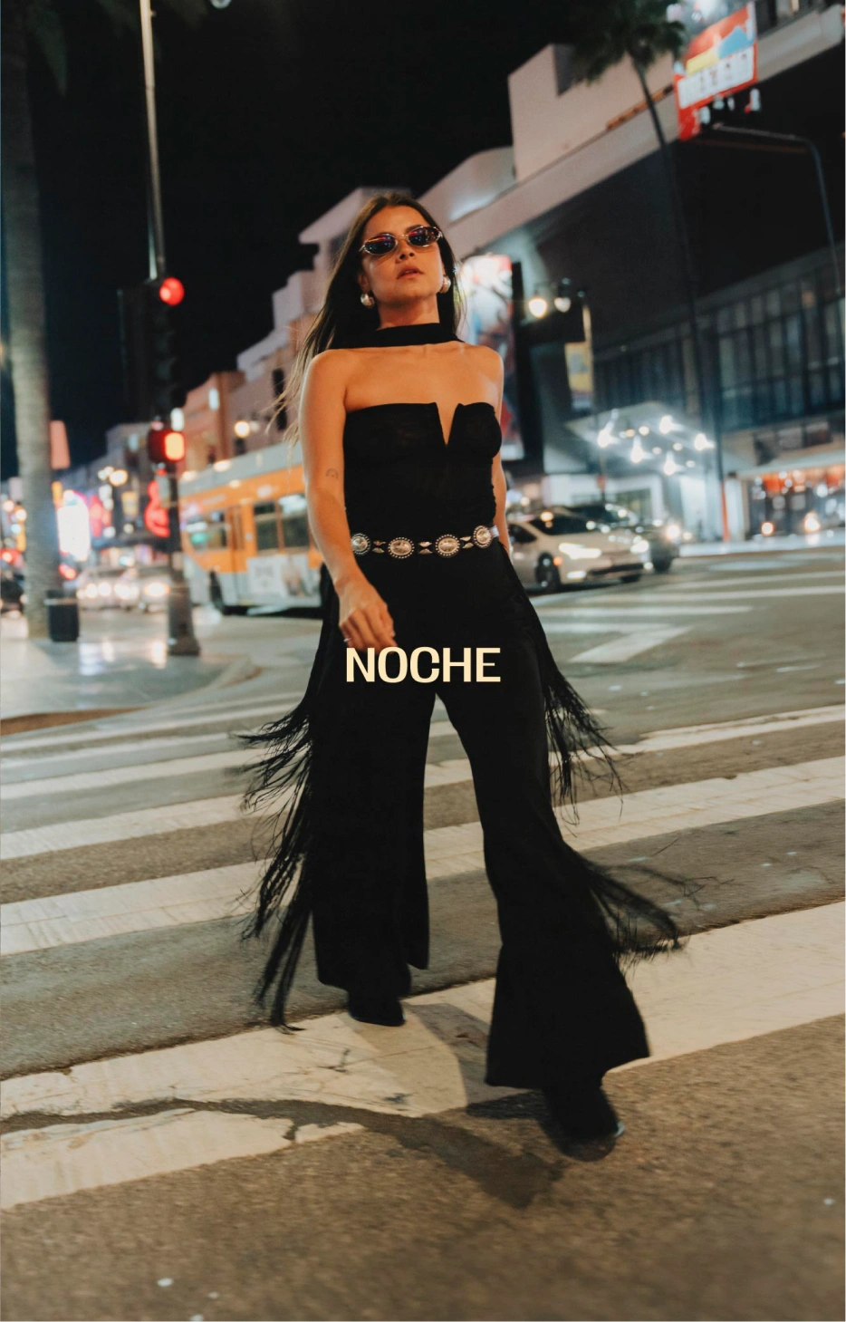 Mujer con enterizo negro strapless, cinturón de herrajes y detalles de flecos en la ciudad de noche.