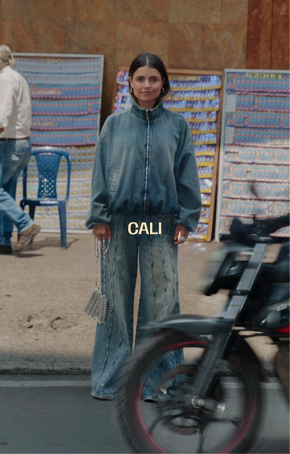 Mujer con chaqueta denim y jeans wide leg en una calle de Cali junto a puestos de lotería.