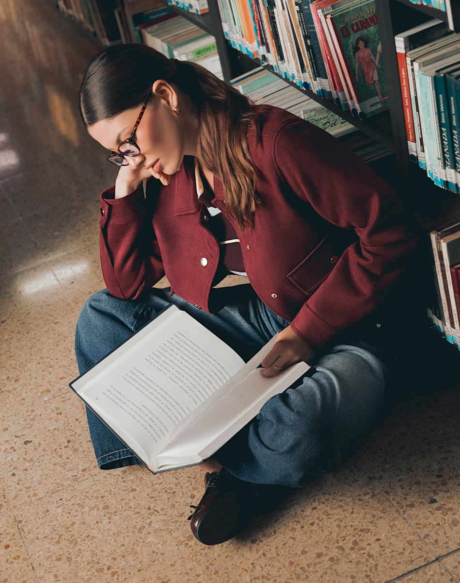 Mujer leyendo y usando chaqueta vino y jeans en el piso; look casual para biblioteca y días de estudio.